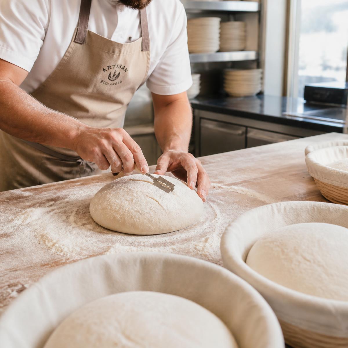 Baker's hands scoring a raw sourdough boule with a lame